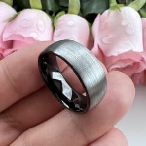 Three fingers holding a plain 8mm contemporary wedding ring featuring a gray brushed finish that extends to its edges and a smooth black sleeve.