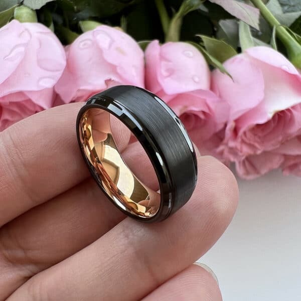 Three fingers bearing an 8mm modern bold wedding ring with a black elevated brushed center, polished edges, and gold-plated sleeve.