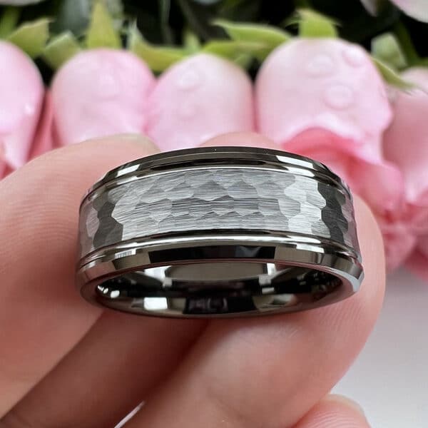 A minimalist men's tungsten wedding band stands on three fingers to show its gunmetal hammered finish and polished beveled edges.
