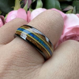 A ring finger wears a black tungsten wedding band featuring polished finish, blue opal center, whiskey wood, and guitar strings.
