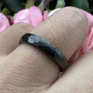 A ring finger wears the 6mm black men's tungsten wedding ring featuring a hammered-brushed center and smooth edges.