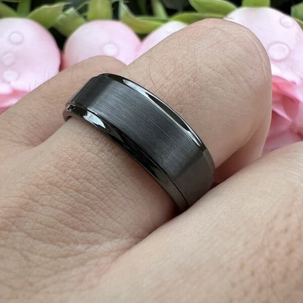 A ring finger wears a 8mm black tungsten men's wedding band with a brushed finish and polished edges.