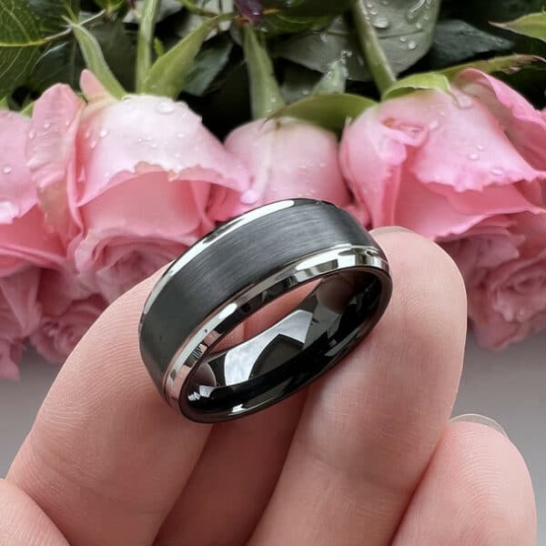 Three fingers tilt a black men's tungsten wedding ring with a brushed center, silver edges, and smooth sleeve on its 9mm width.