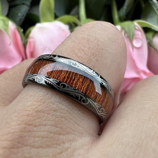 A ring finger wears a black men's tungsten wedding band featuring a KOA wood center, laser etched design, and a polished finish.