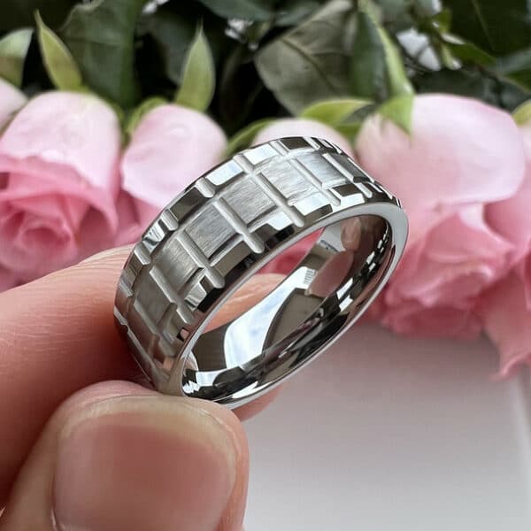 A silver-plated wedding ring with a matrix design, polished edges and sleeve, and brushed center, resting between a thumb and an index finger.