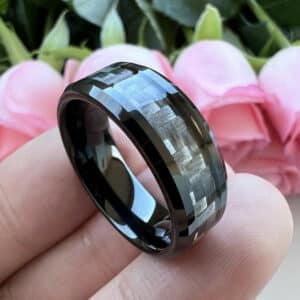 A black men's wedding band with uniquely designed carbon fiber inlay, beveled edges, and polished finish rests on three fingers.