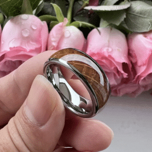 Three fingers holding a wooden wedding band for men featuring polished edges and sleeve, a whiskey barrel oak wood inlay, underneath a domed design.