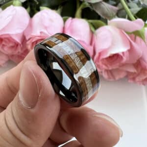 Three fingers hold up a black tungsten men's wedding ring to show its shimmering polished finish over wood and antler inlay.