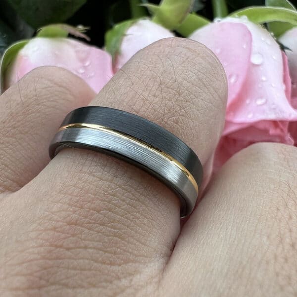 A simple yet stylish wedding band on the ring finger featuring a black brushed finish and a silver brushed finish separated by a gold pinstripe.