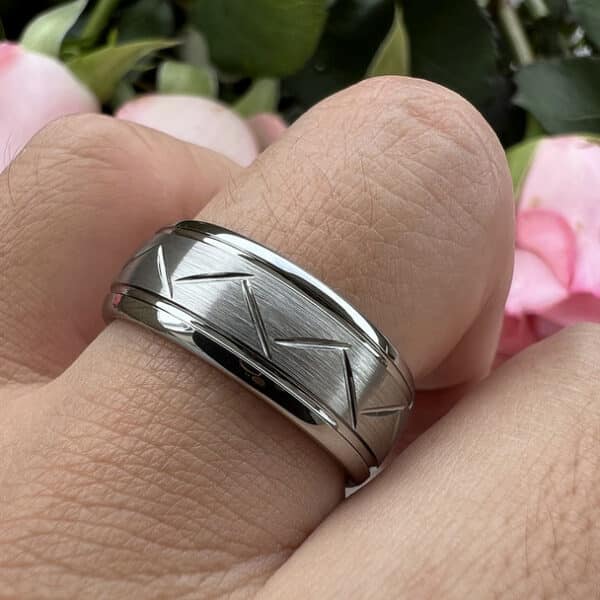 A man's ring finger bearing a minimalist 8mm wedding band with cut cut-through zigzag pattern design, brushed center, and polished edges and sleeve.