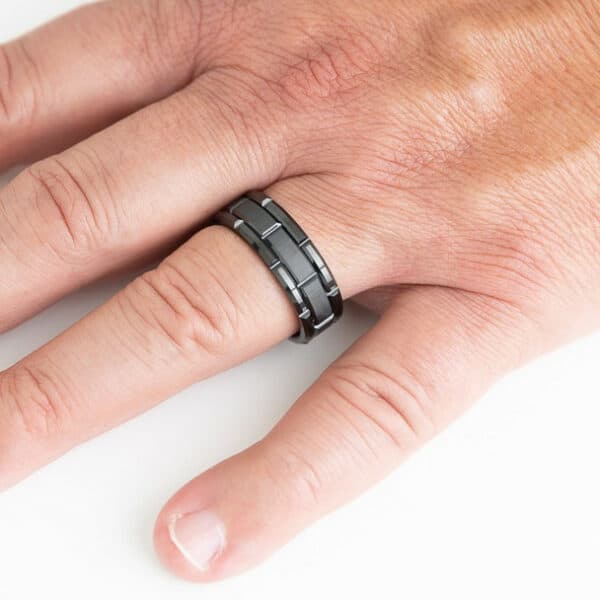 A black and simple wedding band with a carved design, polished edges, and brushed finish on a man's ring finger.