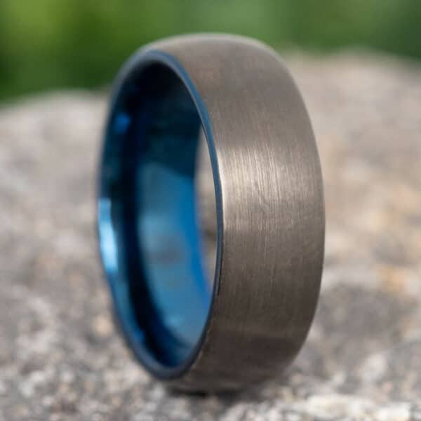 A black tungsten wedding ring for men with a wide 8mm black brushed finish and blue polished sleeve, resting on its curve.