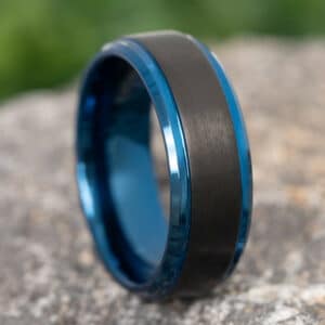 A black tungsten carbide ring with blue sleeves on gravel.