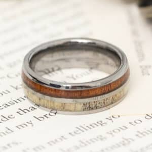 A polished 18k modern white gold wedding band with engraved KOA wood and deer antler resting on a paper surface.