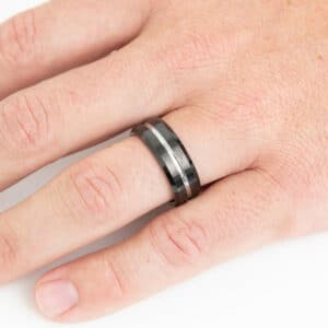 An 8mm black tungsten wedding band worn on a ring finger and features a silver tungsten pinstripe, sleeve, and black sides and edges.