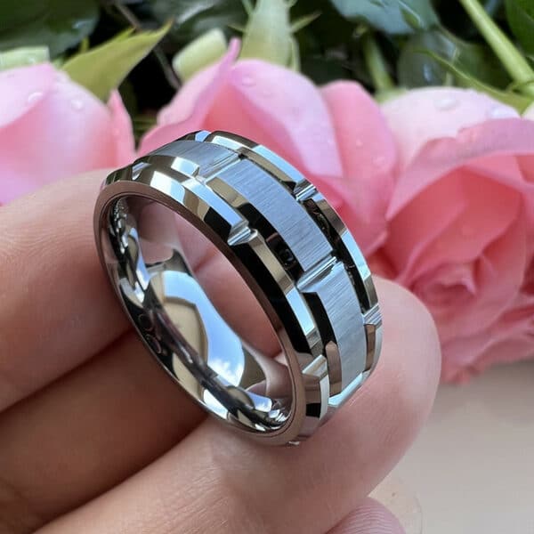 Three fingers bearing a contemporary, industrial-style men's wedding ring with a brushed center, carved pattern design, and polished beveled edges.