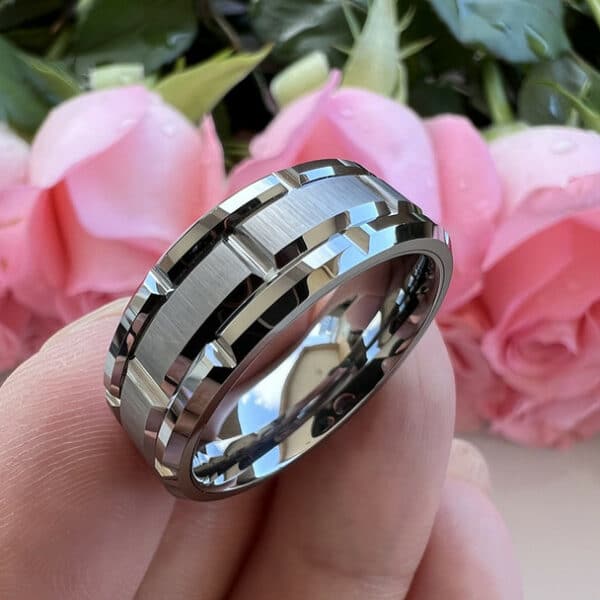 A silver-plated 8mm men's wedding ring with a carved pattern design, brushed center, and polished beveled edges resting diagonally in three fingers.